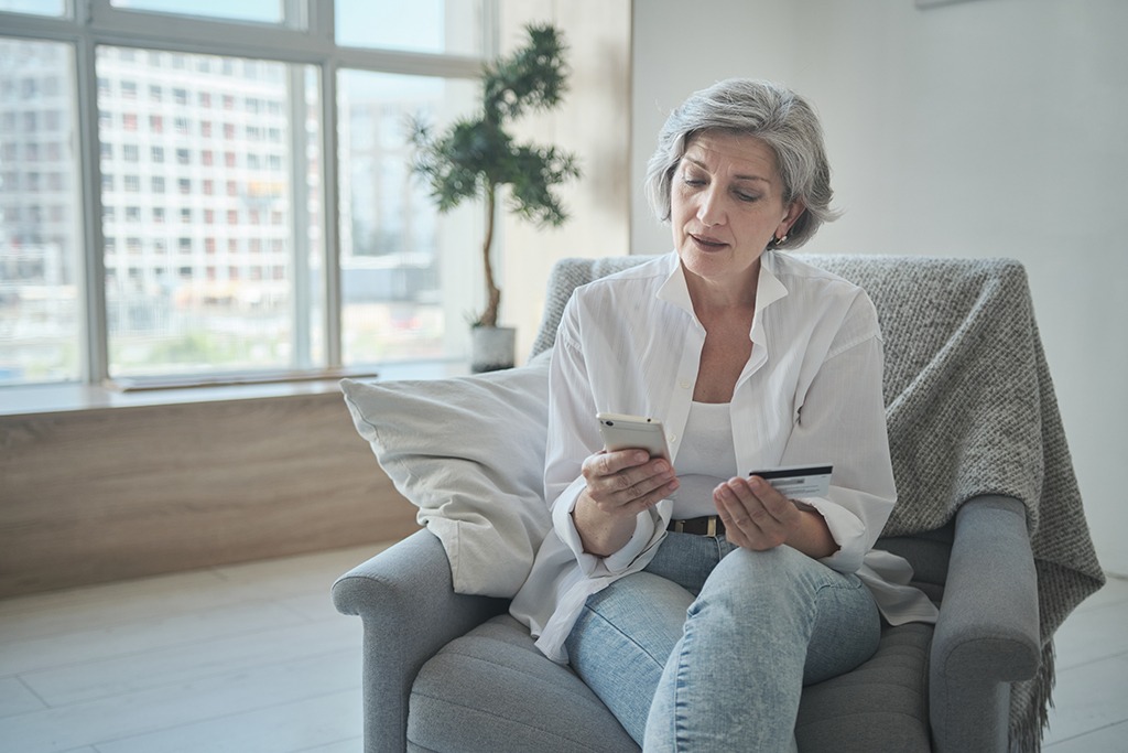 a woman in her 60s sits in a chair, looking at a cell phone