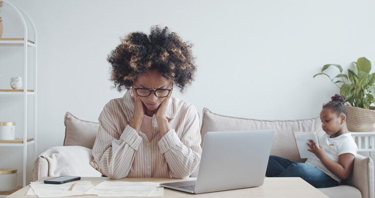 a woman sits at a table with a laptop in front of her, head in her hands, while her child plays behind her