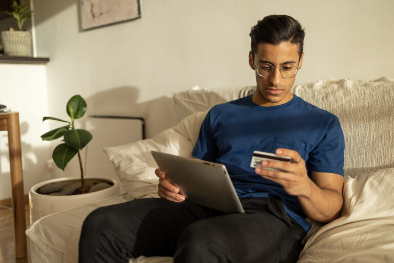 Hispanic man with glasses wearing blue shirt in the apartment using tablet and looking at the credit card