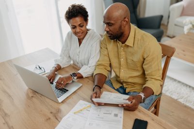 A focused couple is seen working on their budget plans, with one using a laptop and the other reviewing documents on the table. They show determination and cooperation.