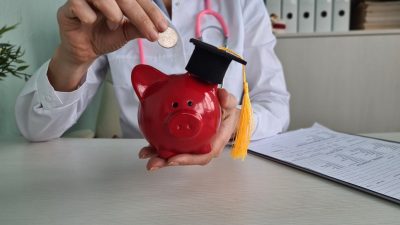 Doctor sitting at table and putting coin in piggy bank. Medical education development and training
