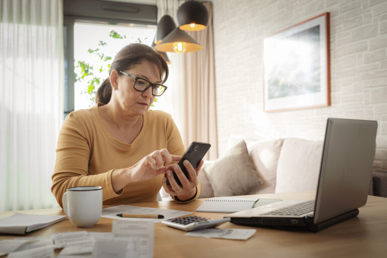 Woman at table with calculator, holding phone thinking about calling debt collector