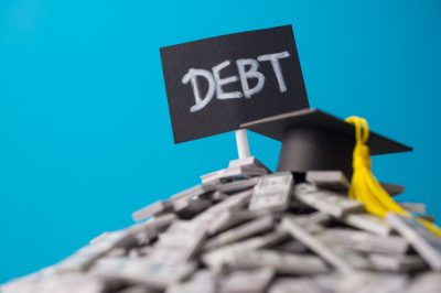 Mortarboard on heap US dollar banknotes and DEBT label with blue background. Student debt or loan, education, studying, investment for future, student financial aid and repayment planning concept.