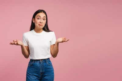 Confused young woman shrugging shoulders, looking at camera posing on pink background