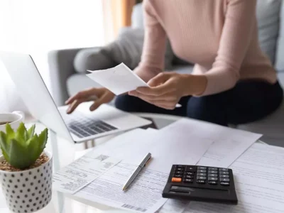 image of a desk scattered with papers, a laptop, and a calculator as a woman types something into the laptop